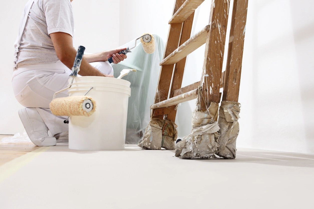 Image of an interior renovation showing a person in the process of painting a wall holding a roller and nearby is a paint bucket and a ladder